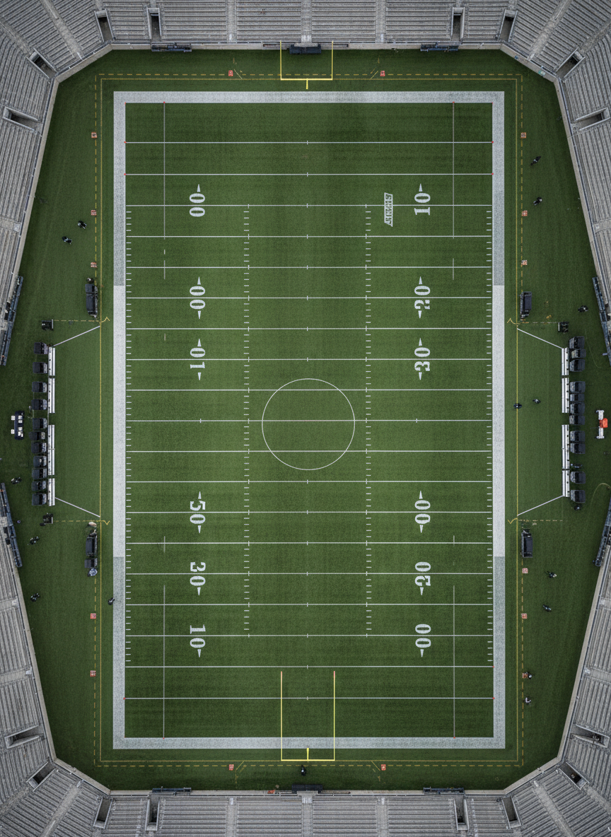 An expansive overhead photographic view of a fully lined NFL field, captured from high above the 50-yard line. Every yard marker, hash mark, and end zone boundary appears crisp against the deep green turf, with subtle wear patterns suggesting a long season of play. The sidelines are neatly arranged with equipment carts, benches, and non-branded gear, but no people. Cool, even overcast daylight spreads across the stadium, minimizing harsh shadows and making the field markings stand out cleanly. The composition is symmetrical, with the midfield logo area intentionally left blank, avoiding any team branding. The mood is objective, data-oriented, and structured, ideal for visualizing schemes, formations, and statistical breakdowns on a professional NFL analysis site.